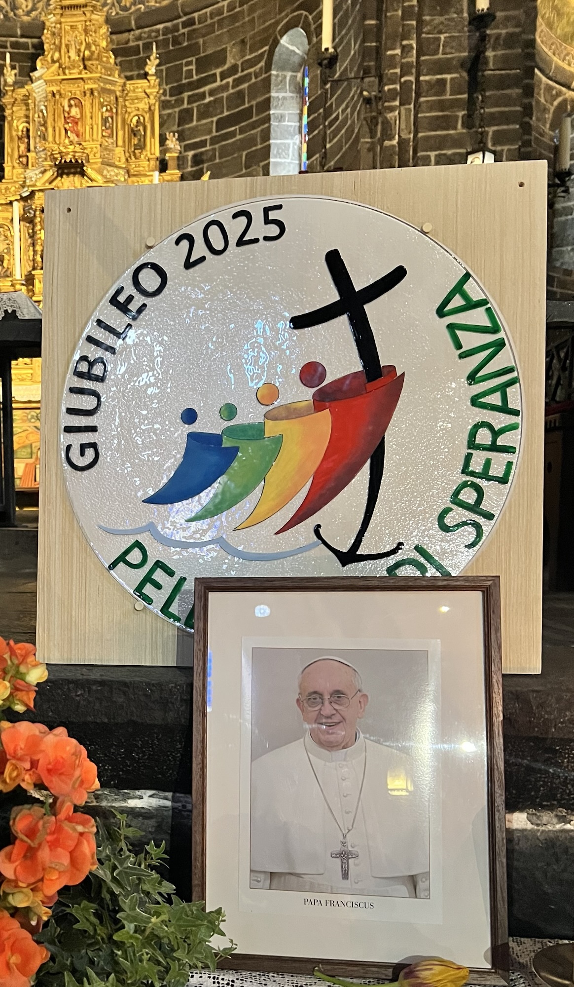 Ein farbenfrohes Schild mit dem Text 'GIUBILEO 2025' und dem Zeichnen von mehreren stilisierten Menschen in Booten, versehen mit einem Kreuz, vor einer festlich dekorierten Kirche. Im Vordergrund steht ein gerahmtes Bild von Papst Franziskus.